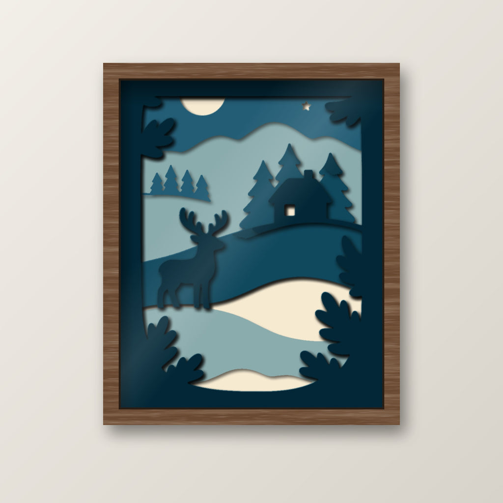 Layered papercut shadow box in deep blues and cream showing a stag standing on a snowy hill in front of a cabin, pine trees, a moon and a star at night.