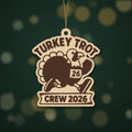 A laser cut wooden ornament showing a cartoon turkey running in a race bib numbered 26, with the words Turkey Trot above and Crew 2026 on a banner below, hanging against a dark green background.