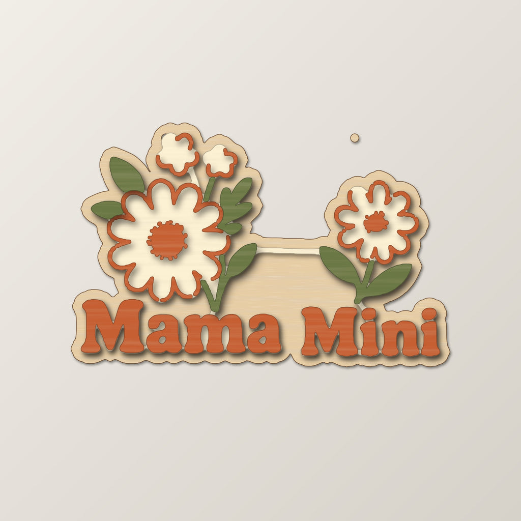 Wooden laser cut ornament with engraved daisy bouquet and single daisy, reading Mama Mini, hanging by a thin cord against a dark green background with golden bokeh lights.